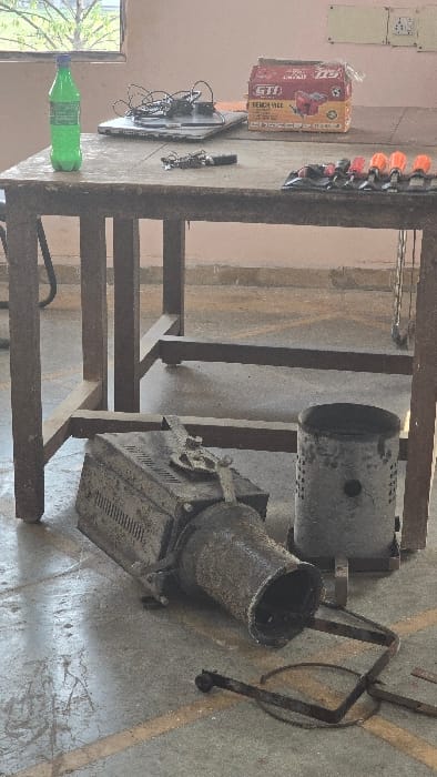 The image captures an indoor scene, likely a workshop or laboratory, featuring a wooden table laden with various items and heavy machinery beneath it. The setting, identified as Gangapur, India, suggests a functional, possibly industrial or educational environment.
On the wooden table, a green plastic bottle, partially identifiable as "SPRITE", stands near a window that reveals outdoor greenery. Next to it, a laptop is open, connected to an array of electronic components and wires, suggesting a prototyping or experimental setup, possibly involving microcontrollers like Arduino. Further along the table, a cardboard box prominently displays "GTI" and "FENCIL CELL" text, with other smaller, unreadable text and a barcode. A black power strip with several orange switches is also visible on the table. The wall behind the table has a light, plain color with electrical outlets mounted.
Beneath the table, two large, robust metallic machines are positioned on a concrete floor marked with yellow lines. One machine is cylindrical, resembling a furnace or a large burner, with perforations and an open top. Adjacent to it is another, more complex piece of equipment with a wide, open top, resembling a mixer or another type of industrial processor, possibly a small foundry or kiln. Its exterior shows signs of heavy use and wear. A rusted metal frame or stand is positioned nearby.
The overall scene suggests a workspace dedicated to hands-on technical or manufacturing activities, possibly involving heating, mixing, or electronic control, but no specific activity is actively taking place. The lighting indicates it is daytime.