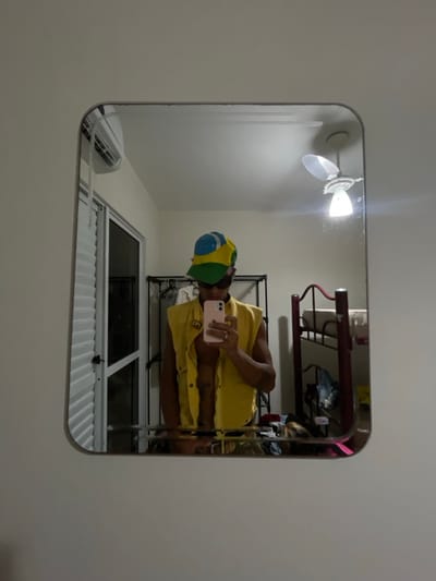 A mirror reflects a man in a yellow shirt and a Brazil-themed hat, holding a pink phone. The reflection includes a ceiling fan, bunk beds with items on the top bunk, a black metal clothing rack, and a window with white corrugated security bars. The mirror hangs against a white wall.