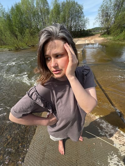 A woman stands on a concrete structure beside a river in Усть-Тунтор, Russia. She wears a gray t-shirt with ruffled sleeves and light-colored shorts. Her hair is brown with gray streaks. She holds her hand to her forehead and looks toward the river. The river flows from left to right in the background, surrounded by trees. The sky is visible at the top left corner.