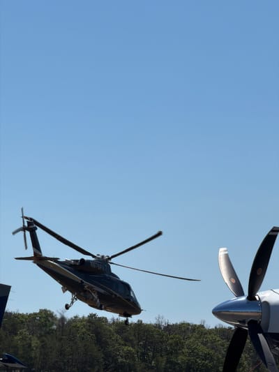 A dark-colored helicopter with registration number N676FX is captured in mid-air, appearing to be either taking off or landing. The helicopter has a sleek design, with gold accents along its body. Its landing gear is extended, suggesting an imminent touchdown or recent lift-off. In the foreground, the prominent propeller of another aircraft, likely a turboprop plane, is visible on the right side of the frame, its blades sharp and metallic. The background features a dense canopy of green trees under a clear, bright blue sky, indicative of a daytime scene. The overall impression is one of aviation activity, possibly at an airport or a private airstrip in a lush, wooded area. The location is specified as the Town of East Hampton, United States of America.