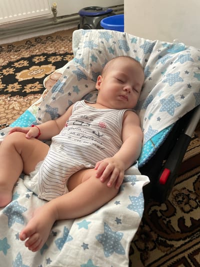 A baby is sleeping peacefully in a portable infant car seat on a patterned rug. The baby is lying on its back, swaddled in a blanket adorned with light blue stars. The baby is wearing a white and blue striped onesie with some writing and a small red emblem. A red string bracelet is visible on the baby's left wrist. The car seat has a black plastic frame and a red latch mechanism. To the left of the car seat, a section of a white radiator and plumbing is visible, suggesting an indoor domestic setting. The patterned rug appears to be in a living room or bedroom. The overall scene is one of quiet domesticity and care. The image has been taken from a high angle, looking down at the sleeping infant.