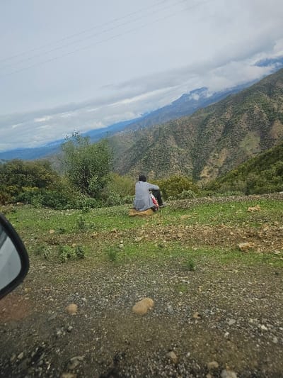 A wide shot depicts a mountainous landscape under an overcast sky, observed from a low vantage point, partially obscured by a vehicle's side mirror in the bottom left. In the middle ground, a lone male figure is seated facing away from the viewer on a low, light-colored object on a patch of green grass. He wears a light-colored long-sleeved garment and dark trousers; a red and white object is positioned near his right side. The immediate foreground consists of a gravelly and stony unpaved surface with sparse green vegetation. The background is dominated by multiple layers of undulating mountains covered in dense green foliage, extending into the distance under a cloudy sky. Distant white structures are visible along the mountain slopes. Several parallel power lines are visible traversing the upper sky. The scene is located in the city of Moqrisset, Morocco.