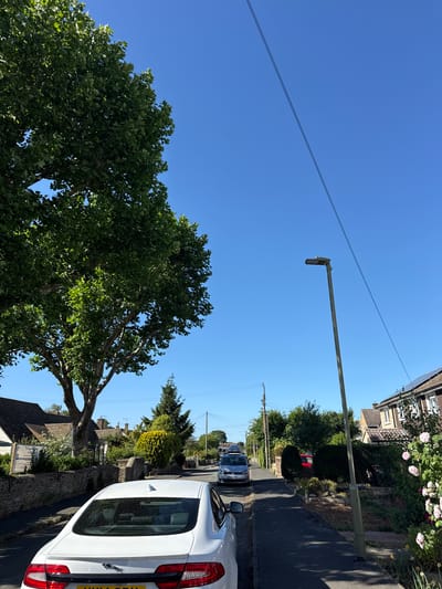 The image captures a street scene in West Oxfordshire, United Kingdom, under a clear blue sky. In the foreground is a large tree with dense green foliage. A white car, partially in the frame on the right, is parked on the roadside. Further down the street, several other cars are visible. A lamp post is present along the side of the road. The road is lined with houses, some with gardens containing greenery and flowers. Power lines stretch across the sky.
