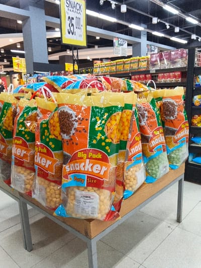 The image captures an indoor scene within a supermarket or grocery store in Sindanglaya, Indonesia, showcasing a large display of snack products. The primary subjects are numerous "Big Pack Snacker Party Size" bags, arranged on a low, light brown display table with metal legs in the foreground.
Each snack bag features a transparent section revealing the product inside, which includes small, round, yellow puffed snacks, brown cereal-like pieces, and green puffed snacks, indicating a variety of flavors or types. The packaging is predominantly bright orange, green, and yellow, with prominent branding "SNACKER" and "Big Pack Party Size." One label on a bag of yellow snacks indicates "NANA" and "1 kg."
In the background, behind the main snack display, are shelves stocked with other packaged goods, including boxes of "ASTOR" wafers and other items labeled "Wafers." Overhead fluorescent lights illuminate the entire scene.
Notable details include several price and informational signs. A large yellow sign above the display reads "SNACK BY NANA 35.000,- 1 PCS," indicating the price for one piece. Other smaller signs with numbers like "20" and "21" are visible further back, likely marking aisles or sections. The overall setting is brightly lit, typical of a retail environment, with a clean tiled floor. The activity depicted is the merchandising of snacks for sale to customers.