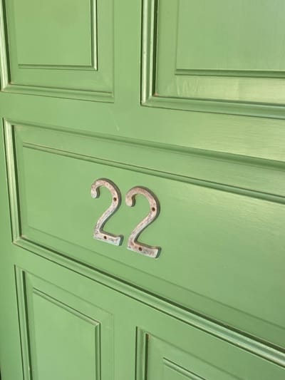 The scene depicts a close-up, angled view of a light green, paneled door, likely located in Πλατύς Γιαλός, Greece. Centrally affixed to one of the door panels are two metallic house numbers, "22". The numbers appear to be made of a light-toned, possibly weathered or patinated metal, each secured by two small, dark screws. The door surface exhibits rectangular recessed panels and a smooth, painted finish. Minor surface imperfections are visible, including a faint vertical streak above and to the left of the numbers. No people, actions, or specific environmental context beyond the door are visible.