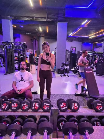 The image depicts the interior of a modern gym facility, identified as located within the Municipal Unit of Corinth, Greece. The foreground is dominated by a two-tiered metal rack holding numerous black rubber hex dumbbells, with visible weights including 10KG and 12KG, featuring the brand "OKO" (or "OKAE" mirrored).

In the midground, a woman stands centrally, facing a large mirror and holding a black smartphone to take a mirror selfie. She has long brown hair, wears a dark athletic top and matching dark leggings, with a tattoo visible on her upper left arm. To her left, a man is seated on a black weight bench, looking down at a white smartphone he holds. He has short dark hair and a beard, wearing a white t-shirt with a black "Jumpman" logo and dark red athletic pants. To the right, another man walks away from the camera, wearing a light-colored t-shirt, blue shorts, and blue athletic shoes, holding a dark object, possibly a water bottle.

The background reveals a spacious gym environment equipped with various fitness machines, including cable machines and weight racks, under a distinctive lighting scheme featuring blue and orange/yellow LED strips along the ceiling and walls. The floor is a dark, speckled rubberized surface. Mirrored walls reflect the gym's interior and additional indistinct patrons.