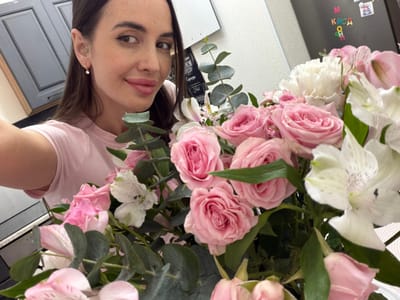 The image captures a close-up of a woman holding a large, colorful bouquet of flowers, taken from a slightly upward angle.
In the lower right portion of the frame, a woman is visible, looking up and slightly towards the left. She has long, dark brown hair that falls over her shoulders and a gentle, pleasant expression with a subtle smile. She is wearing a light pink t-shirt, and a small, light-colored stud earring is visible in her right ear. Her lips appear full and are either naturally colored or have a very light pink tint.
The majority of the image, especially the upper and left sections, is dominated by a vibrant bouquet. The most prominent flowers are numerous light pink roses, showcased in various stages of bloom, from tight buds to more open, flourishing petals. Interspersed among the roses are several white alstroemeria (Peruvian lilies), which feature delicate petals with distinctive dark speckles on their inner surfaces. Towards the lower left of the bouquet, some ruffled white flowers, possibly carnations, can also be seen. The bouquet is enriched with abundant green foliage, including broad, silvery-green leaves, likely eucalyptus, and darker green leaves associated with the roses, adding texture and depth.
In the background, to the far left, a dark surface, possibly a refrigerator, is visible, adorned with several colorful magnets. Clearly decipherable text on one magnet shows the Cyrillic letters "К И С А" (KISA) arranged vertically, and another magnet with the letter "М" (M) further below. To the right of the woman, a dark, paneled door or cabinet with a visible frame is present in the background. The lighting is bright and even, suggesting an indoor setting with ample illumination. The image conveys a sense of happiness and appreciation, likely capturing a moment of enjoying the beautiful floral arrangement.