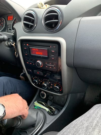The image shows the interior of a car. A person's hand, wearing a watch and a ring, is on the gear shift. Sunglasses and a key fob are visible in the center console. The car's radio is on, displaying "FM1-P2 NOVOE". The dashboard includes air vents and instrument clusters. The environment appears to be a city in Belarus, possibly Курапольскі сельскі Савет. The image is taken from the perspective of a passenger or the driver.
