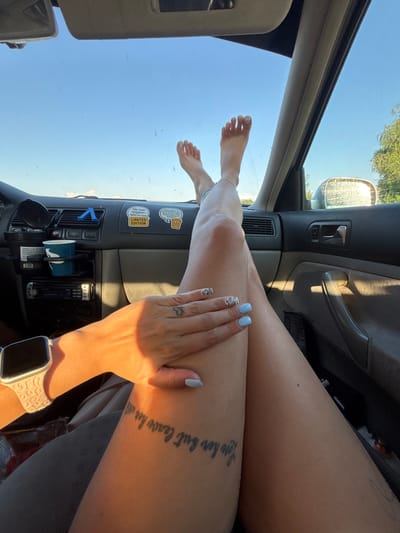 An individual's legs are positioned in the passenger seat of a car, with their feet resting on the dashboard. The exterior environment is visible through the windshield and side window, indicating a sunny day with a clear blue sky and distant trees. The car's interior is partially visible, showing the dashboard with various items including a car stereo, air vents, cup holders, and stickers. A hand with painted nails is resting on the thigh, and a tattoo is visible on the leg. The scene suggests a casual moment during a car journey. The location is identified as Dolni Bogrov, Bulgaria.