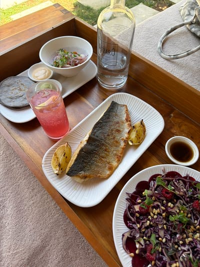 This image shows a meal served on a wooden tray. The tray holds a bottle of water with the brand "ATZARÓ IBIZA" visible, a plate of grilled fish with lemon wedges, a small bowl of dark sauce, and a salad with red cabbage, raspberries, and pine nuts. To the left, there are small bowls containing what appears to be ceviche or a similar dish, a creamy sauce, and a pink beverage with ice and lemon. There are also grey corn tortillas on a small white plate. A beige patterned bag with handles rests on a light-colored surface in the background. The setting appears to be outdoors, with hints of greenery and a textured, possibly towel-like surface beneath the tray, suggesting a relaxed, possibly resort-like environment. The lighting and composition suggest a bright, daytime setting. The overall impression is one of a well-presented meal, likely enjoyed in a leisurely manner, possibly at a beachside or poolside location given the Ibizan context. The visible text confirms the location as Ibiza, a Spanish island known for its vibrant atmosphere and beautiful scenery.
