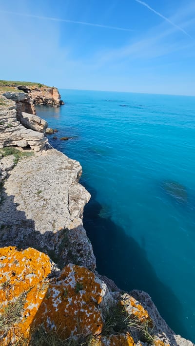 The image captures a scene from Kamen Bryag, Bulgaria. The viewpoint is from a cliff overlooking the Black Sea. The water is a vibrant turquoise, contrasting with the rocky coastline. The cliffs are composed of layered rock formations, some covered with orange lichen and sparse vegetation. The sky is clear blue. In the distance, along the coast, there are more cliff formations and a built structure that could be part of an old fort or staircase. There is no visible human presence in the scene.