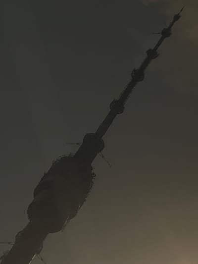The image is a low-angle, silhouette shot of the Ostankino Tower in Moscow, Russia, against a hazy, overcast sky. The tower, a prominent broadcast and observation tower, is shown from its base upwards, diagonally across the frame. The structure is dark and appears to be made of metal. Multiple levels of platforms or structural elements are visible along its length. The sky is a uniform, muted gray with a slightly brighter area towards the top right, possibly indicating the direction of the sun or a break in the clouds. The overall impression is one of scale and imposing height, with the tower dominating the frame. The specific location in Vëshki, Russia, is incorrect as the Ostankino Tower is located within Moscow city limits.