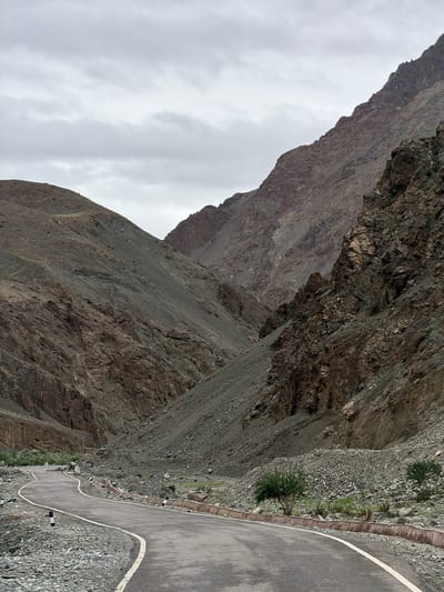The image shows a winding asphalt road with white lines cutting through a mountainous, arid landscape. The mountains on either side are steep and rocky, with varied shades of brown and gray. The sky is overcast with a thick layer of gray clouds. Along the side of the road, there are small, sparse bushes and rocks. A few white and black bollards line the edge of the road. The scene appears to be a remote, mountainous region, likely a highway or a road leading through a pass. The presence of a road and bollards suggests human infrastructure in an otherwise natural and rugged environment. The image is taken from a low angle, looking ahead along the curve of the road. The overall impression is one of vastness and a sense of journey.