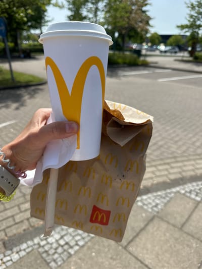 A person holds a McDonald's cup with a golden arches logo and a brown paper bag with multiple golden arches and a red McDonald's logo, likely containing food. A wrapped straw is protruding from the bag. The person is wearing a wristwatch with colorful bands. The background shows a parking area with cars, trees, and a paved walkway. The location is likely in or near Ottersberg, Germany, as inferred from the general context of McDonald's locations.