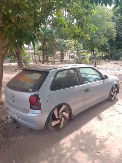 In Cáceres, Brazil, a silver Volkswagen Gol, a compact hatchback, is parked on a dirt road beneath the shade of trees. The car, identified by the license plate "MJD-6175," displays custom modifications, including aftermarket wheels with a complex, turbine-like design and a red "Deluxe" sticker on the rear. A yellow decal reading "Deixe" is affixed to the dusty rear windshield. The vehicle appears weathered, with visible dust accumulation on its surfaces and a slightly lowered stance. The surrounding environment is rural or semi-rural, characterized by dry dirt, scattered leaves, and dense green foliage from the surrounding trees. In the background, glimpses of residential structures and other vehicles, including what appears to be a dark SUV, can be observed. The lighting suggests a sunny day, with dappled shadows cast by the trees across the car and the ground.