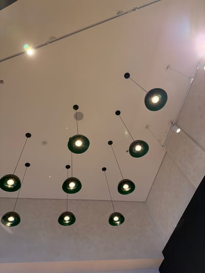 The image shows a ceiling with several modern pendant lights. These lights consist of dark green spherical shades with visible bulbs. Each light is connected by a thin black wire to a small, circular black fixture mounted on the ceiling. The lights are arranged in a non-linear pattern across the off-white ceiling. There are also track lighting fixtures visible on the ceiling. The top right of the image shows a contrasting dark, almost black, section of the ceiling or a portion of the building's architecture. The lights appear to be indoors. The location of the scene is unknown.