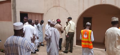 The image depicts a group of men standing outside a building with a pale, textured wall and an arched entryway. The scene appears to be outdoors, with natural lighting suggesting it is daytime. The weather seems to be warm, as many of the individuals are wearing light clothing and traditional attire such as robes and caps.

The subjects are primarily men, most of whom are wearing traditional Nigerian clothing. Several individuals are wearing caps of various designs. Two men in the foreground and middle ground are wearing uniforms that include vests over their clothing. One vest is bright orange with reflective stripes and features a portrait of a man and text. The text on the vest reads "KADUNA STATE TASK FORCE". Another man is wearing a light-colored uniform with a cap. The group appears to be gathered and conversing, with some gesturing with their hands. One man in the middle of the frame is smiling.

The setting appears to be in an urban or semi-urban area in Nigeria, possibly Sabon Gari North, given the context. The building's architecture suggests a warm climate. The overall mood seems to be one of a gathering or a meeting, with a generally positive and engaged atmosphere. There are no obvious objects beyond the clothing and the building itself, which has a closed door and a window visible through the archway.