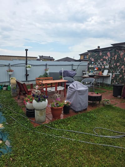 The image depicts a backyard scene in Проводник, Russia, featuring a patio area with outdoor furniture and decorative elements. The patio is constructed with red brick pavers and is surrounded by a green lawn.
In the center of the patio, there is a wooden picnic table with benches. To the left of the table, a red wooden chair is placed. Several potted plants, including petunias and other flowering species, are arranged around the patio. Two large, white woven baskets hold plants, and a brown clay pot sits near them.
To the right of the patio, a gray tarp covers an object, possibly a grill or outdoor equipment. Adjacent to this is a circular fire pit with a metal grate. Further to the right, a tall black trash can is visible.
Along the back of the patio, a gray metal fence runs horizontally. Hanging from the fence are several white planters with pink and white flowers. Behind the fence, houses and other structures can be seen, along with a cloudy sky overhead.
A trellis covered in a floral pattern is attached to a structure on the far right, adding a decorative touch to the area. Various gardening tools, such as watering cans, are located on the left side of the image, near the lawn. The scene appears to be set up for outdoor relaxation and entertainment.
