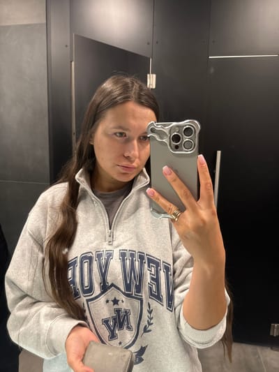 The image displays a mirror selfie taken by a young person, likely female, indoors. The scene is rotated 90 degrees clockwise, suggesting it was captured with the phone held horizontally and then uploaded.
The person has long, dark brown hair that falls over their shoulders and chest. They are wearing a light gray pullover or sweatshirt with a half-zip neckline. The sweatshirt features a prominent dark blue graphic print on the front. Above a shield or crest design, the word "NEW" is clearly visible, followed by letters that appear to spell "WEIL" or "WELL". Inside the crest, the text "ROY" is legible, and below it, two stars contain the letters "Y" and "M" respectively.
The person's face is visible, looking towards the camera (phone). They have dark eyes and a neutral expression. Their right hand holds a smartphone, which is dark gray or silver-colored and encased in a distinctive clear or metallic-looking case with wavy, "melted" edges, particularly around the triple camera module. The person's nails have a French manicure style with pink bases and white tips. Rings, possibly three simple bands, are visible on a finger of their right hand. In their left hand, they hold a small, light beige or gray wallet or cardholder, which appears somewhat worn.
The background, visible in the mirror reflection, consists of dark-colored panels or walls, possibly black or dark gray, with visible seams or joints creating horizontal lines. A silver-colored handle or bar is also reflected on the dark surface. The setting appears to be a functional indoor space, such as a restroom or changing area.