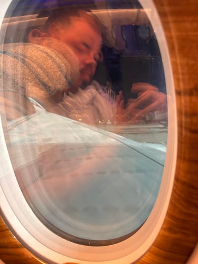 An oval window with a light wood frame encases a blurred scene. The reflection shows a person sleeping, holding a mobile phone. Outside, a blurred airplane wing is visible. The lighting is warm, possibly from the cabin lights, casting a reddish hue on the sleeping figure. The overall image is distorted due to movement and reflection in the window.