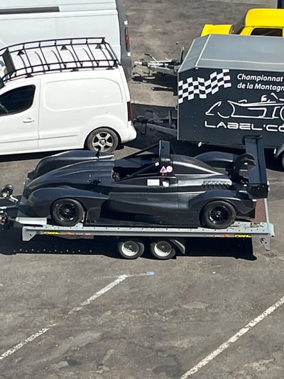 In Espalion, France, a black, open-wheel race car is parked on a trailer on a paved surface. The car has a sleek, aerodynamic design with a prominent rear wing and exposed wheels. It is positioned on a silver-colored trailer with multiple wheels, suggesting it is being transported. In the background, a white van with a roof rack is visible, along with a portion of a black trailer featuring checkered flag graphics and text that reads "Championnat de la Montagne" and "LABEL'CO". A yellow vehicle is also partially visible behind the black trailer. The scene appears to be an outdoor event or a staging area for a race or automotive gathering.