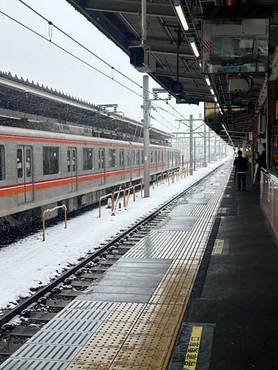 A long shot looking down a train platform covered in fresh snow. A silver and orange train occupies the left side of the frame. Yellow tactile paving edges the right side of the platform. A person stands near the end of the platform in the distance. Above the train, a station building with electrical wiring and support structures is visible. It appears to be snowing heavily.