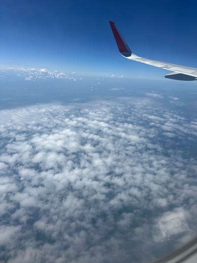 The image shows a view from an airplane window, looking out at a vast expanse of sky and clouds. The airplane's wing, painted white with a red tip, is visible in the foreground on the left side of the frame, extending diagonally upwards. Below the wing, the sky transitions from a deep, clear blue near the horizon to a lighter, hazy blue. A dense layer of fluffy, white cumulus clouds stretches across the majority of the frame, appearing textured and uneven. The sun is likely high in the sky, as indicated by the bright illumination of the clouds and the clear visibility, suggesting it is daytime. The overall impression is one of a serene flight, offering a glimpse of the world from above. There is no visible text in the image. The location context suggests the flight is over or near Шульгин Лог, Russia.