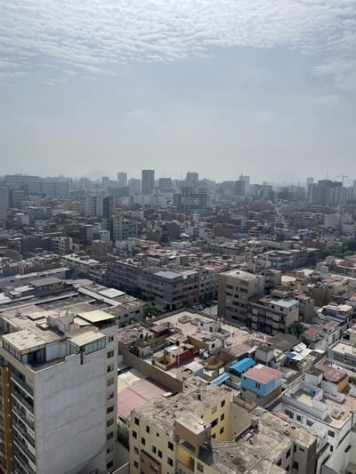 An aerial, slightly angled, medium shot captures a dense urban landscape in Lince, Peru, characterized by a sprawling array of buildings under a hazy sky. The sky is a pale blue with scattered, wispy white clouds. In the distance, a slightly obscured skyline of taller, modern buildings is visible, suggesting a more developed area beyond the immediate foreground. The mid-ground and foreground are dominated by a multitude of multi-story buildings with flat roofs, packed closely together. These structures exhibit a variety of architectural styles and color palettes, ranging from off-white and beige to shades of gray, brown, and some with red accents. Many rooftops are covered with various structures, including water tanks, antennas, and what appear to be informal rooftop additions or extensions made from corrugated metal and plastic sheeting in blue and red. Some buildings have visible balconies, and in the lower left, a taller, modern building with balconies is prominent. Evidence of human activity is minimal, with no people clearly visible. The overall impression is of a densely populated, established urban environment with visible signs of organic growth and development.