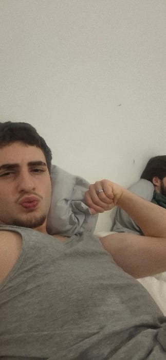Two men are lying down. One is facing the camera with pursed lips and a wink. He is wearing a gray t-shirt and a ring on his left hand. His head rests on a gray pillow. The other man is blurred in the background, appearing to be asleep. A white wall is partially visible to the left. The photo appears to be taken from a slightly rotated angle.
