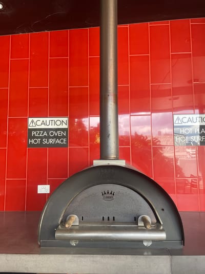 The image shows a close-up of a pizza oven in a commercial setting. The oven is dark grey and arched, with a metal flue pipe extending vertically upwards. Two wooden handles protrude from the front of the oven. On the red tiled wall behind the oven, there are two cautionary signs. The sign on the left reads "CAUTION PIZZA OVEN HOT SURFACE." The sign on the right is partially obscured but appears to read "CAUTION HOT PLAT HOT SURF." Below the left sign, a white electrical outlet with three sockets is visible on the red tiled wall. The overall environment appears to be an outdoor or semi-outdoor area, possibly a restaurant patio or food stall, given the red tiled backsplash and the type of equipment. The image does not provide enough context to determine its specific location within the city of Brisbane, Australia. There are no people visible in the frame.