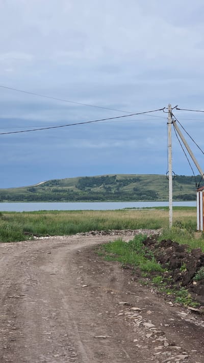 The image depicts a rural landscape near Kupoyarovo, Russia, dominated by a dirt road in the foreground leading towards a body of water. A power pole with visible wires stands on the right side of the road, adjacent to a small, partially visible structure with white and reddish-brown siding. Beyond the road and a strip of tall grass, a wide, calm body of water stretches across the midground. In the background, a rolling, verdant hill covered in trees and patches of grass rises against a cloudy, overcast sky. The overall scene suggests a quiet, natural setting.