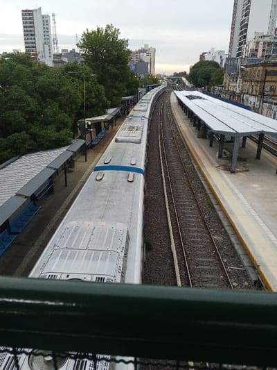The image provides an elevated, matter-of-fact view of the Ramos Mejía train station in Argentina. A multi-car, silver-colored train with blue accents on its roof is stationary on the leftmost track. This track is adjacent to a platform equipped with a dark gray corrugated roof structure and blue benches. To the train's right, an empty set of tracks parallels the train, followed by a second, elevated concrete platform, mostly covered by a lighter gray corrugated roof structure. Multiple tracks are visible, curving slightly to the right in the distance. The urban background comprises a mix of tall residential and commercial buildings, some appearing modern, others exhibiting signs of age and weathering, interspersed with dense green foliage. The sky is overcast, suggesting diffused light conditions. No individuals are discernible on the platforms or within the immediate train vicinity. A dark green metal railing occupies the lower foreground, indicating the elevated perspective from which the photograph was taken.
