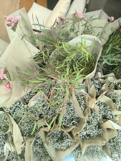 The image shows a collection of floral arrangements wrapped in newspaper print paper. The flowers are a mix of small white flowers, pink flowers, and eucalyptus greenery. The arrangements are densely packed together. The environment appears to be a flower shop or market. Based on the presence of "Leidschendam" printed on the paper wrapping the flowers, it is inferred that this photo was taken at a flower shop/market in Leidschendam, Netherlands.