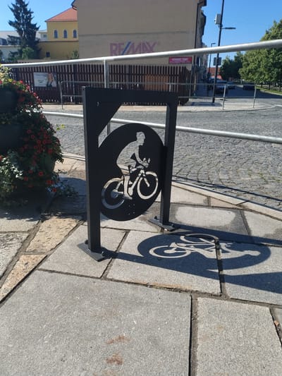 In Písek, Czechia, a black metal bicycle rack with a silhouette of a cyclist is positioned on a paved walkway. The rack is adorned with a circular cutout featuring the cyclist, whose shadow is cast onto the ground. To the left, a large planter overflows with red flowers and green foliage. In the background, a wooden fence separates the walkway from a street lined with trees and buildings. A sign with "RE/MAX" branding is visible on one of the buildings. The scene is illuminated by bright sunlight, indicating daytime.
