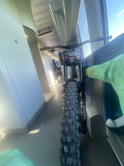 The image shows a front view of a mountain bike inside a train carriage. The bicycle is predominantly featured in the foreground, with its front tire, handlebars, and part of the frame clearly visible. A person wearing a bright green jacket is partially visible on the right side of the frame, their arm extended towards the bike. The background reveals the interior of the train, with seats, windows showing daylight, and a visible sign indicating a toilet ("WC") on the left. The scene appears to be set in Merichleri, Bulgaria, within the confines of a moving train.
