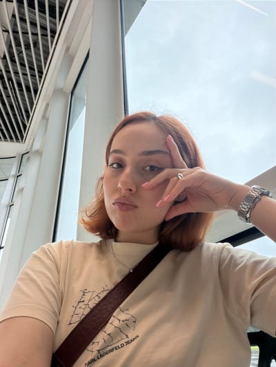 A young woman with auburn hair is posing for a selfie. She is wearing a light beige t-shirt with a print, a silver watch, and a silver ring. Her nails are painted light pink. She has a brown leather strap across her body. The background includes a bright sky, a window, and a ventilation grill. The location appears to be indoors, potentially a building with modern architecture. There is no information to define the city of Ianeti, Georgia.