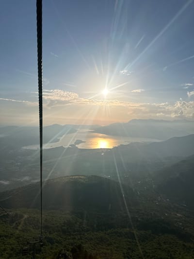 An aerial view captures the sun setting over a body of water, likely the Bay of Kotor, in Čavori, Montenegro. The sun, positioned in the upper center of the frame, casts bright rays across the sky, illuminating the water's surface with a golden sheen. In the foreground, a thick, dark cable descends vertically, suggesting the perspective is from a cable car. The landscape is mountainous, with rolling hills covered in dense green forests and dotted with some scattered settlements visible on the slopes. Distant mountains are silhouetted against the hazy sky. The overall atmosphere is serene, with the warm light of sunset creating a dramatic and picturesque scene.