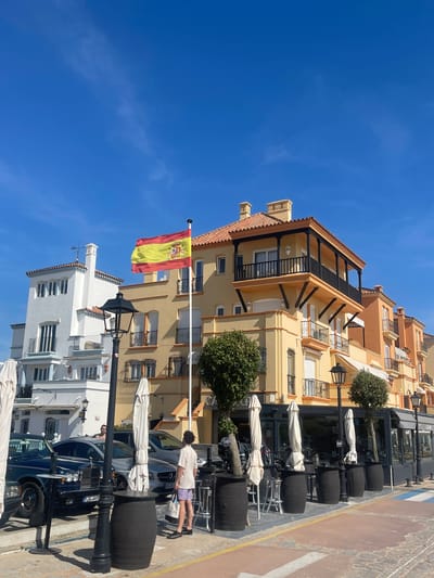 The image shows a street scene in El Puerto de Santa María, Spain. The foreground features a row of barrels likely repurposed as tables or decor for a restaurant or café. Umbrellas are folded and positioned near these barrels. Several cars are parked along the street. A person is walking between the vehicles and the barrel arrangement. The Spanish flag is prominently displayed from a pole attached to a building. The buildings in the background are of varying heights and architectural styles, with one white building contrasted against a multi-story yellow building with balconies. The sky is clear and blue.
