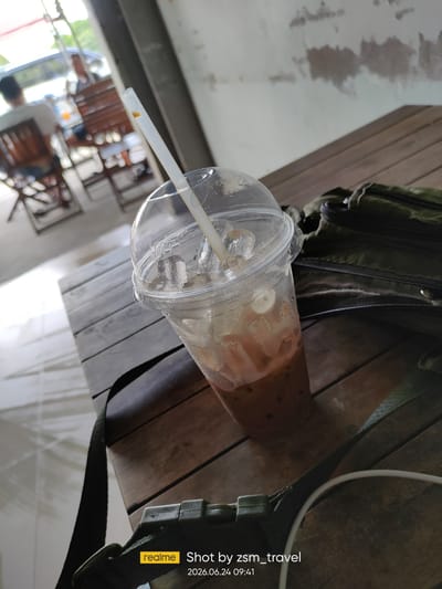 A close-up shot focuses on a clear plastic cup filled with an iced coffee drink, resting on a wooden table. The cup has a clear domed lid with a white straw inserted, and is partially filled with ice cubes and a brown liquid that appears to be coffee mixed with milk or cream. Condensation has formed on the outside of the cup. Behind the cup, a dark green bag with zippers is visible. In the background, out of focus, are several wooden chairs and tables, suggesting an outdoor seating area of a cafe or restaurant. The floor appears to be tiled. A watermark at the bottom of the image reads "realme Shot by zsm_travel 2026.06.24 09:41". The overall scene suggests a casual moment at a cafe in Ben Cau, Vietnam.