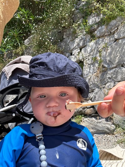 A young child, estimated to be a toddler, with fair skin and blue eyes, is centered in the frame, looking forward. The child wears a navy blue bucket hat and a blue long-sleeved rash guard featuring a white logo on the chest. A grey beaded pacifier clip is attached to the rash guard. Dark brown ice cream or chocolate is smeared around the child's mouth and drips onto the chin and rash guard. From the right, an adult's hand offers a small wooden spoon containing a scoop of light-colored ice cream topped with a dark brown substance. To the left, a portion of a grey stroller with a beige blanket draped over it is visible. The foreground consists of rocky and pebbly ground. The background is dominated by a stone wall covered with green foliage and small white flowers. A part of a straw hat is visible in the upper left corner. This outdoor scene is located in Abbadia Lariana, Italy.