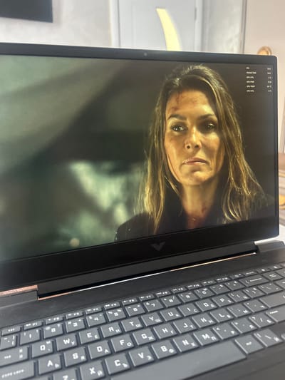 The image is of a laptop screen displaying a scene of a woman with messy hair and what appears to be blood or dirt on her face and neck. The laptop's keyboard is visible in the foreground, with keys in a Cyrillic script. On the top left of the laptop screen, system performance statistics are displayed. The surrounding environment appears to be indoors, with a blurry background suggesting a room interior. There is no information to suggest this image was taken in Topolovgrad, Bulgaria.