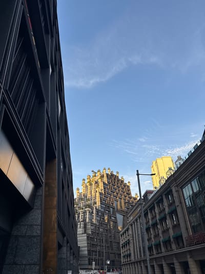 The image captures an upward perspective of modern city architecture set against a clear blue sky. In the foreground, spanning the bottom left of the frame, a dark, contemporary building rises with a distinct faceted design, featuring angular lines and panels that appear to be dark stone or metal, with subtle reflective bronze accents near its base.

To the right, and extending upwards from the mid-ground, a striking golden-toned building stands out. It is characterized by multiple staggered levels and balconies, many of which are adorned with green foliage, creating a visually rich, terraced effect. Further into the background on the upper right, other tall urban structures are partially visible, including one with numerous windows and what appear to be green awnings or shading elements. A lamppost or street light is positioned towards the upper right, extending into the frame.

The sky is a vibrant, clear blue with delicate, wispy white clouds scattered across it, indicating fair weather. The scene is brightly lit by natural sunlight, suggesting either morning or late afternoon, as the light illuminates the golden building and casts directional shadows on the dark foreground structure. No human activity or visible text is present, rendering the image a detailed study of architectural forms and their interplay with the natural light and sky in an urban environment.
