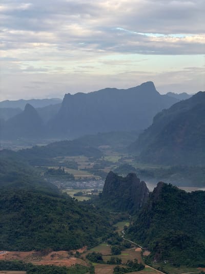 The image captures a panoramic view of a mountainous landscape, likely in or around Hong Ngern Village, Laos. The foreground features steep, densely forested green hills with some patches of cultivated fields and a visible dirt road leading up a reddish-brown cleared area. The midground showcases dramatic, jagged limestone karst peaks jutting out of verdant valleys, with a winding road and what appear to be rice paddies. Further in the distance, successive layers of hazy blue mountains recede into the background, softened by atmospheric perspective. The sky above is overcast with soft, horizontal clouds in shades of grey and white, with hints of pale blue peeking through. No people or discernible activity are visible in the scene. The overall impression is one of natural grandeur and rural tranquility.