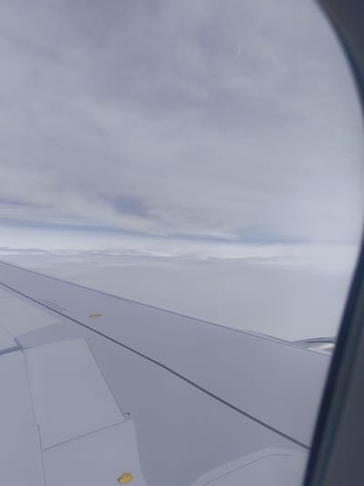 The image is an aerial view from an airplane window, looking out over a vast expanse of clouds. The airplane wing is visible in the foreground, spanning across the bottom half of the frame. The wing is a light grey color and appears smooth, with some panel lines and warning labels visible. There are also two yellow markings on the wing, possibly emergency exit indicators.

Above the wing, a thick layer of clouds stretches to the horizon, appearing as a soft, white blanket. The sky above the clouds is a pale blue, with some wispy clouds and a hint of hazy sunlight. A thin, crescent-shaped object, likely a very thin cloud formation or an artifact of the camera lens, is visible in the upper left portion of the sky.

The overall scene is serene and suggests a flight high above the earth. There are no indications of any specific landmarks or features that would pinpoint the location in Xã Phú Hòa, Vietnam, or any other city. The image focuses on the atmospheric conditions and the perspective from an aircraft.