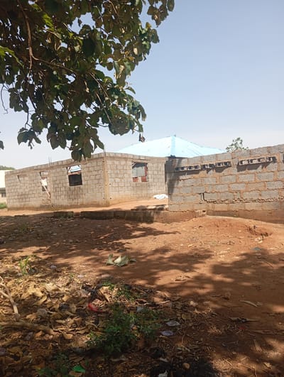 A partially constructed building with cinder block walls stands under a clear blue sky in Karsana 2, Nigeria. A light blue corrugated metal roof covers a section of the structure. In the foreground, dry leaves and debris litter the red dirt ground, with a leafy tree branch extending from the upper left corner. The building appears to be a single-story structure, with openings for windows and doorways visible. A higher, unfinished wall of cinder blocks is situated to the right of the main building.