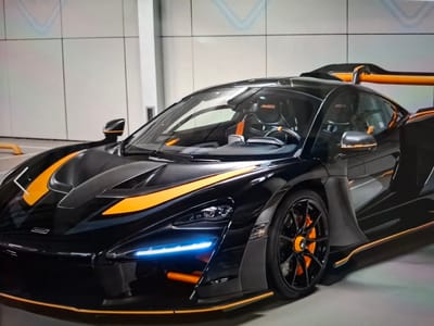 A black McLaren Senna with orange accents is parked indoors, likely in a garage or showroom. The car is positioned at an angle, showcasing its aerodynamic design. The exterior features sharp lines, a prominent rear wing, and distinctive orange racing stripes along the hood and side. The headlights emit a bright blue glow. The interior, visible through the tinted windows, includes bucket seats with "SENNA" branding in orange. The background consists of a light-colored, tiled wall and a gray floor. The overall impression is one of a high-performance, modern sports car. There is no information within the image to suggest it is located in Argun, Russia.