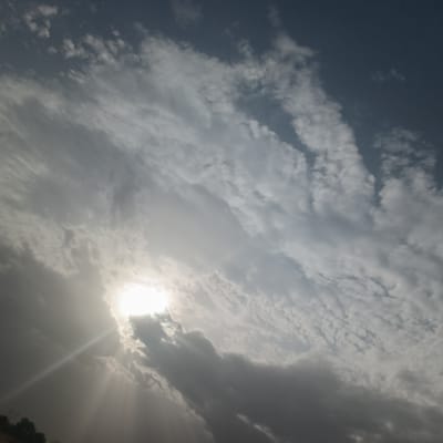 The image shows a daytime sky with a bright sun partially obscured by clouds. The clouds are a mix of altocumulus and darker storm clouds. Sunlight is streaming through gaps in the clouds. The image is rotated counterclockwise. There is no information to identify the specific location in Kariya, Nigeria, and there are no people or objects other than the sky and clouds visible.
