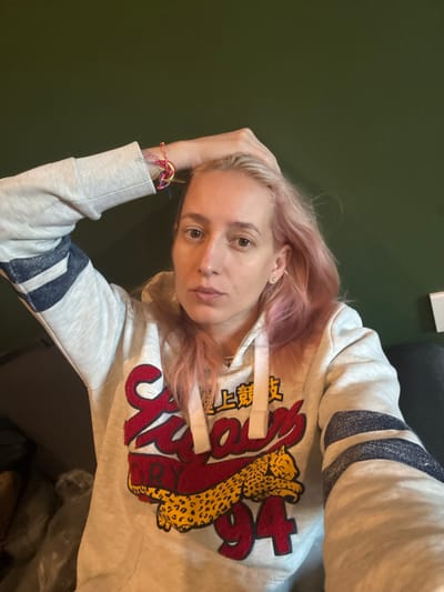 A woman with light pink hair rests her head on her hand, wearing a grey hoodie with navy stripes on the sleeves and a stylized graphic of a leopard and the number 94 in red. The background is a solid dark green color, except for a black object visible in the top right corner. She looks directly at the camera with a slightly serious expression. A colorful bracelet is visible on her wrist.