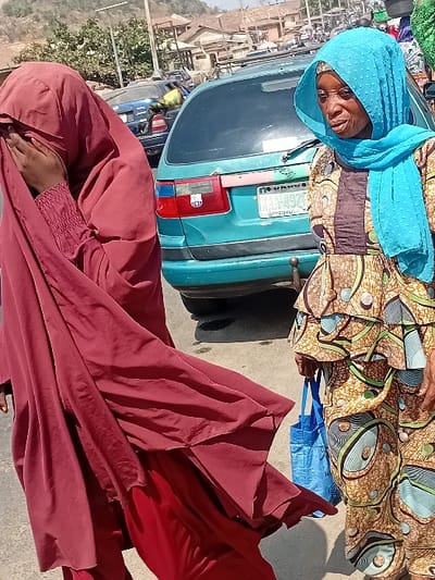 Two women stand outdoors. One, wearing a light blue headscarf and patterned brown and yellow clothing, stands behind a light blue car. Her expression is smiling. The other woman, draped in a flowing dark red garment, partially obscures her face. The background shows a sunny outdoor scene with hints of buildings and distant foliage.