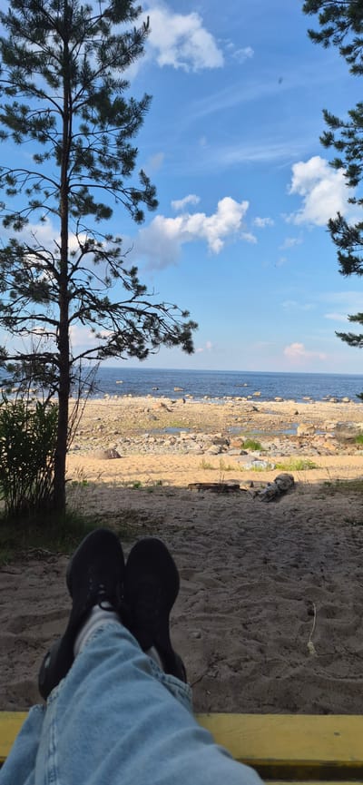 This is a photo taken from a low angle, looking up towards a blue sky with white clouds and framed by the branches of a pine tree. The scene transitions to a rocky beach with calm blue water beyond. In the foreground, a person's legs, clad in blue jeans and black shoes, are visible, resting on a yellow surface, likely a bench. The sand is a light brown, with some dry grass growing in patches. The overall impression is one of a peaceful day at the beach, possibly in the afternoon given the lighting. There is no visible text in the image.